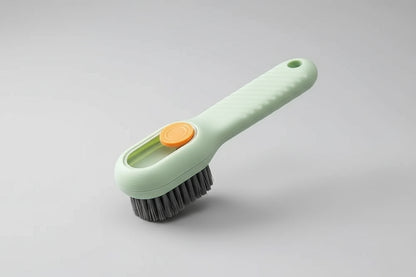 Hand using a green cleaning brush with an orange button to scrub a white sneaker on a beige background.