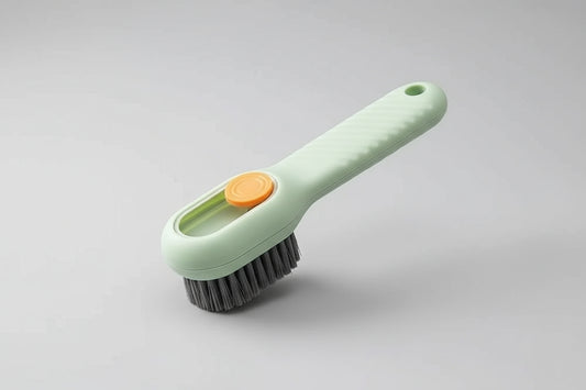 Hand using a green cleaning brush with an orange button to scrub a white sneaker on a beige background.